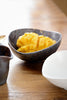 Close-up of the Charcoal Grill Shell Sharing Dish holding cubed mango — textured charcoal glaze with caramel splatter detail