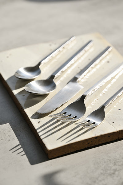 Square-handled brushed stainless steel cutlery set arranged on a handmade ceramic platter with directional sunlight