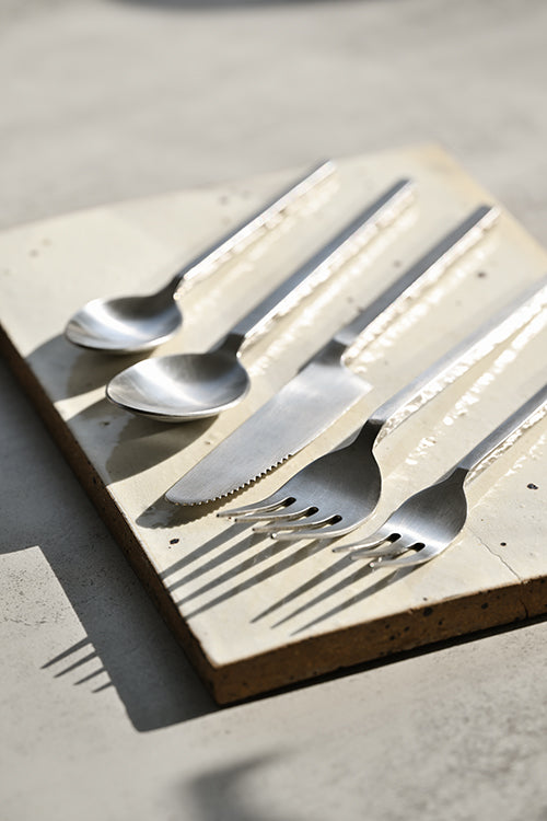 Square-handled brushed stainless steel cutlery set arranged on a handmade ceramic platter with directional sunlight