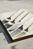 Square-handled brushed stainless steel cutlery set arranged on a handmade ceramic platter with directional sunlight