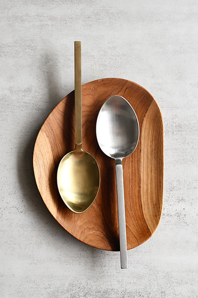 Square-handled brushed stainless steel and brushed gold serving spoons side by side on a wooden tray, top view