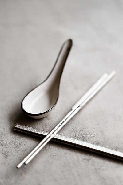 Square-handled brushed stainless steel chopsticks paired with a handmade ceramic soup spoon on a textured concrete surface
