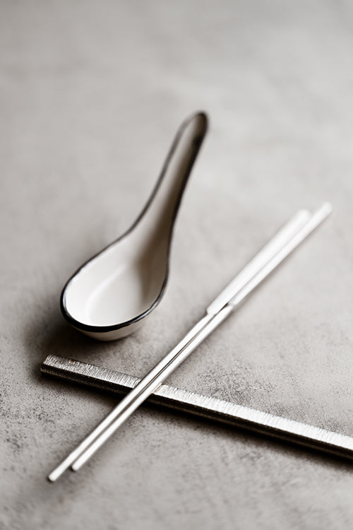 Square-handled brushed stainless steel chopsticks paired with a handmade ceramic soup spoon on a textured concrete surface