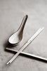 Square-handled brushed stainless steel chopsticks paired with a handmade ceramic soup spoon on a textured concrete surface